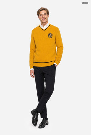 A man modeling a mustard yellow V-neck sweater with black and white trim and a crest on the chest, worn over a white collared shirt with black trousers and black shoes, against a light gray background.