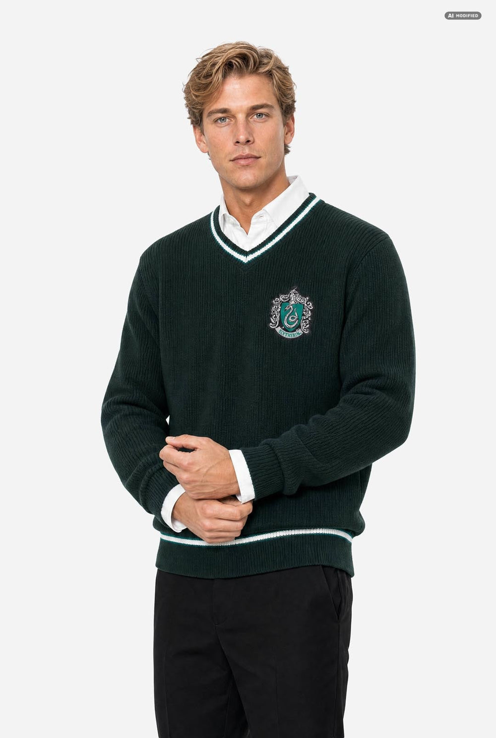 A man wearing a dark green knit v-neck Harry Potter sweater with white and light green trim, featuring a Slytherin crest on the chest, layered over a white collared shirt and paired with black trousers, set against a plain light gray background.