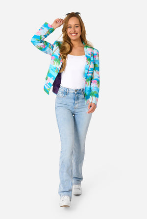 Image with a light background and a woman wearing a light blue blazer with tropical flamingo print and jeans. Holding sunglasses on her head. Full body image. 