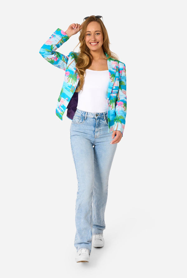 Image with a light background and a woman wearing a light blue blazer with tropical flamingo print and jeans. Holding sunglasses on her head. Full body image. 