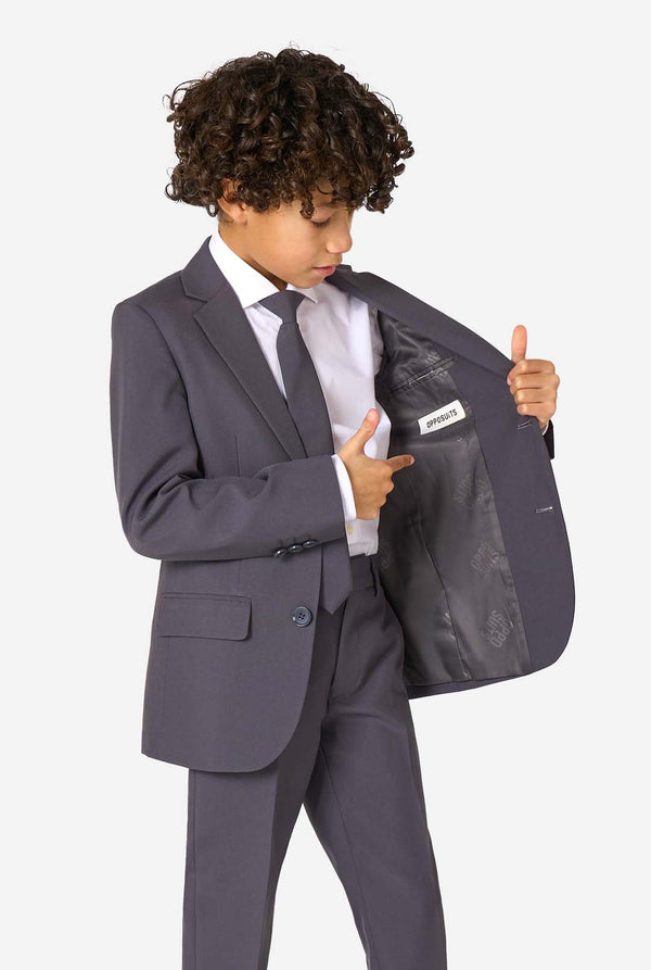Child wearing a gray suit with a white shirt and black tie, holding open the jacket to show a label.